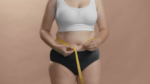 Plus size caucasian female in white bra and black briefs measuring her waist with measure tape for control weight. Studio still no head anonymous shot video. 