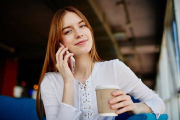 Portrait of attractive hipster girl with takeaway caffeine beverage resting in cafeteria and using public internet connection for mobility calling, young female with coffee to go posing while talking