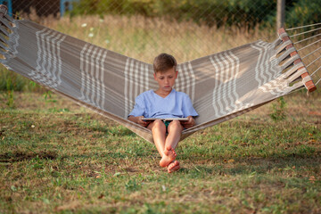 Cheerful little boy reading book lying on the hammock