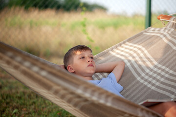 Cute child boy reading book and dreamink on hammock in backyard on summer day. Kid relaxing in hammock.