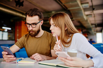 Millennial man and woman with smartphone devices browsing website discussing collaborative learning during brainstorming meeting in coffee shop, happy students with cellphones talking at table