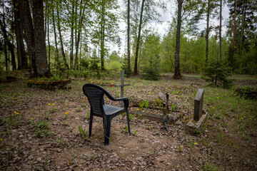 a plastic chair in old Latvian forest park cemetery. Trees, grass, sand, metal catolic cross, gravestone