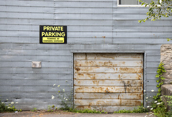 Side of building with grungy bent and rusted corrugated grey metal siding and dented and rusted white garage door, private parking sign, weeds, nobody	

