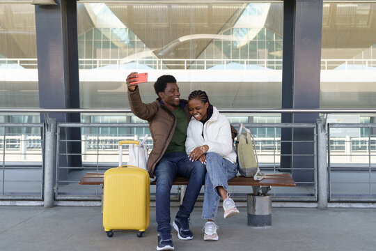 Happy laughing couple of tourists taking selfie outdoors airport terminal wait for bus or taxi after arrival to vacation, loving black man and woman travel. Safe tourism after end of covid-19 concept