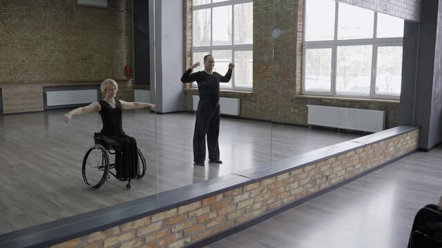 Professional middle aged dancers, man and handicapped woman on wheelchair doing warm up exercises in front of mirror in ballroom. Disabled female and her dance partner during training