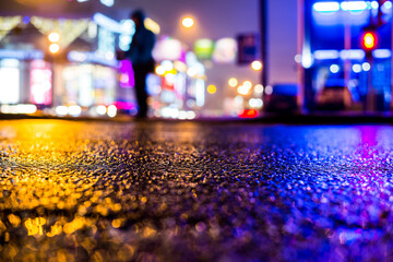 Rainy night in the big city, illumination lights of the shopping center with a man standing on the road. Close up view from the asphalt level