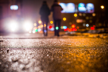 Rainy night in the big city, glare from the headlights of the car parked and people passing near. Close up view from the sidewalk level