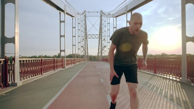 Young runner injured his knee during training. Morning running on the pedestrian bridge in the city at dawn. High quality 4k footage