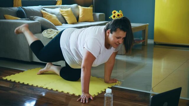 Overweight young woman kneels on a mat and raises her legs while her network personal trainer talks by video call