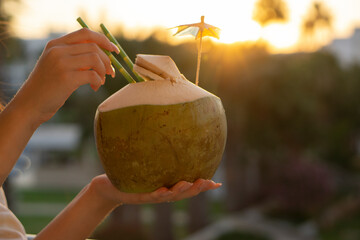  Woman hands  holding  fresh  green coconut drink with paper straw  and rainbow umbrella   on tropical  sunset background with copy space . Vacation  exotic  travel destinations  concept . - Powered by Adobe