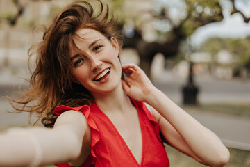 Optimistic woman in red dress making selfie outside. Happy girl with sloppy brown wavy hair in bright outfit smiling outdoors..