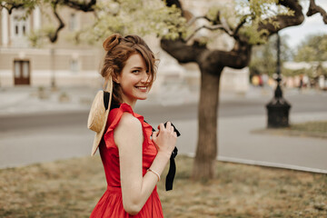 Charming girl with briwn hair in bright outfit smiling in park. Happy woman with cool hat in red dress looking into camera outside..