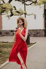 Obraz premium Bright girl in red summer dress posing at street. Beautiful woman with brown hair putting modern hat and looking away outdoors..