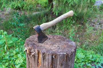 A small old ax with a wooden handle is stuck in a wooden block with a damaged surface from use. A green forest is visible in the background. Background.