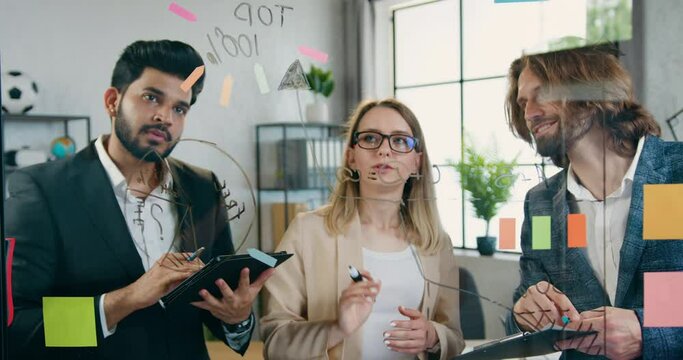 Work in office of three ingenious young professional people over new start up project in modern office with reminders on glass wall and sticky notes