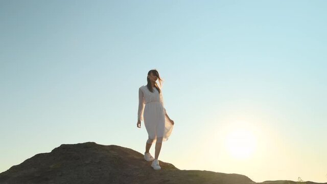 beautiful slender woman in translucent white dress and white sneakers descending from top of hill, sun shines into camera, sunset or dawn. young lady walks through mountains with sky in background