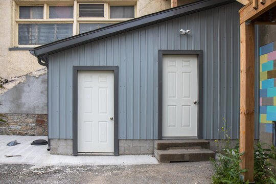 White metal entrance doors on building annex with sloped roof, corrugated grey metal siding in front of stucco building with barred windows, nobody