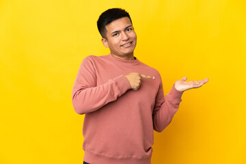 Young Ecuadorian man isolated on yellow background holding copyspace imaginary on the palm to insert an ad
