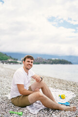 Young smiling man sitting on bedspread on pebble beach with childrens toys