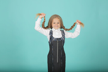 a beautiful little smiling blonde girl in a white shirt and a gray sundress a schoolgirl holds her pigtails in her hands