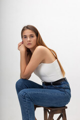 young attractive caucasian woman with long brown hair in corset, blue jeans on white studio background. skinny pretty lady posing with bare feet on wooden chair or ladder. portrait of beautiful female
