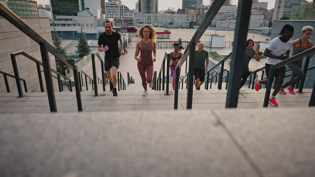 Team group training. Young men and women, a mixed race company, dressed in jerseys and shoes, run up the outdoor stairs in a large modern city