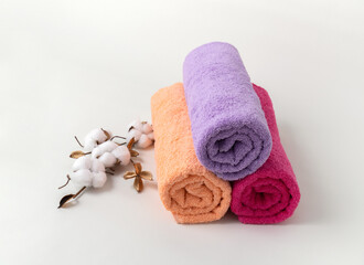 Large colored cotton terry towels for the body close-up on a light background with a cotton branch. Lilac, peach and burgundy towels, rolled into rolls. Concept: natural textile bath accessories.