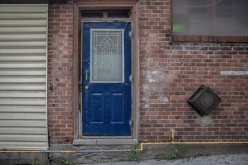 Exterior wall of building with weathered red brick, dented blue metal entrance door with glass insert, horizontal vinyl siding, gas pipes, gas vent, graffiti, nobody