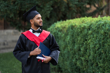 Portrait of handsome indian graduate student in graduation glow with diploma looking forward for...