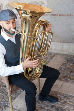 A jazz musician dressed in vintage style, playing the tuba on the street in broad daylight. With beard and goatee dyed blue and a retro cap.