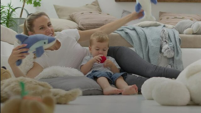 Happy and smiling mom with baby playing in living room at home with plush stuffed animals toys, sitting on the floor with pillows, caucasian blonde with blue eyes, healthy and love life growth concept