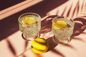 Two glasses of water with sliced lemon on sunlit background with shade. Summer refreshment concept. Minimal style.