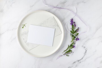 Floral wedding invitation card mockup. Blank paper postcard, vintage envelope on white plate and bunch lavender flowers with ribbon on light marble background. Top view with copy space