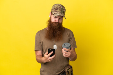 Fototapeta premium Redhead Military man with dog tag isolated on yellow background holding coffee to take away and a mobile