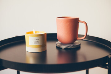 Cozy interior details. Little round table with ceramic cup of delicious tea and yellow aromatic candle. Resting after busy work day, relaxing concept. Selective focus.