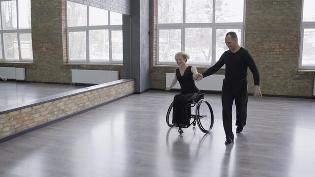 Cheerful ball dancing couple having fun in dance hall, woman with impaired mobility laughing while dance partner driving and circling her in wheelchair. Joyful dancers fooling around in ballroom