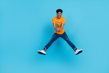 Fototapeta na wymiar Full size photo of hooray brunet millennial guy jump with fruit wear orange t-shirt jeans isolated on blue color background