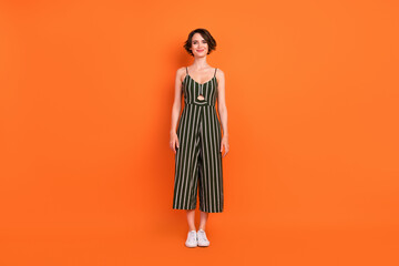 Full size photo of nice brunette young lady stand wear overall sneakers isolated on orange background
