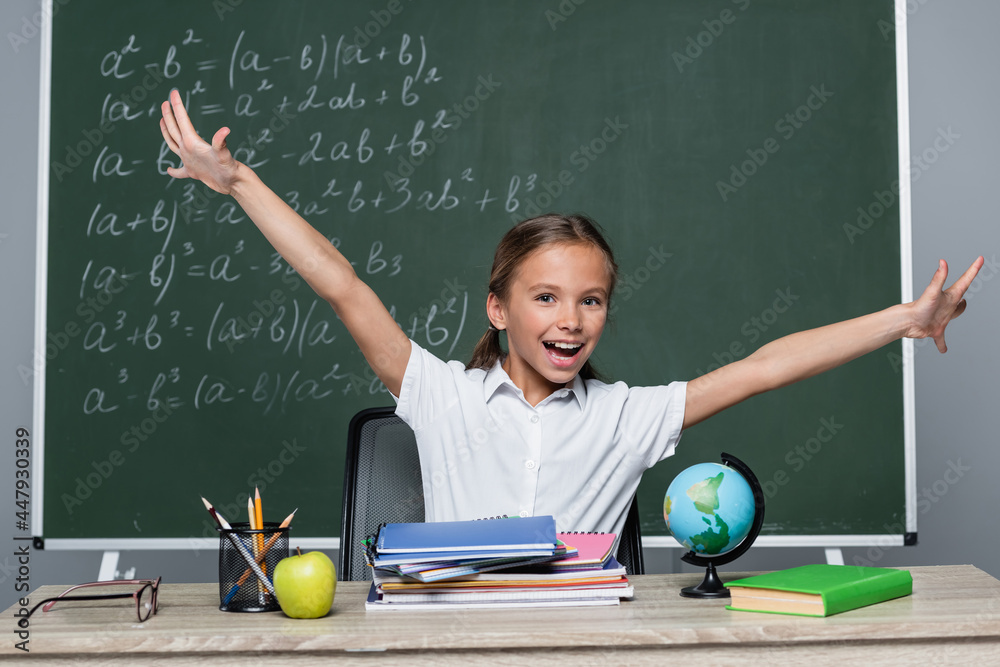 Wall mural cheerful schoolgirl with open arms near notebooks on desk and chalkboard with equations