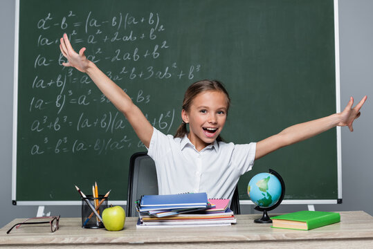 cheerful schoolgirl with open arms near notebooks on desk and chalkboard with equations