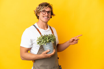 Gardener blonde man holding a plant isolated on yellow background pointing finger to the side
