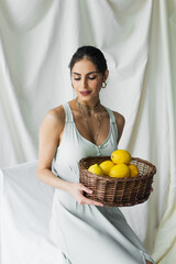 Obraz premium happy woman in dress holding wicker basket with lemons on white