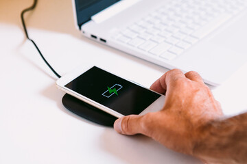 Hand with white mobile smart phone with the logo of a battery with green lightning on the screen, charging on a wireless charger base next to a laptop on a white work table