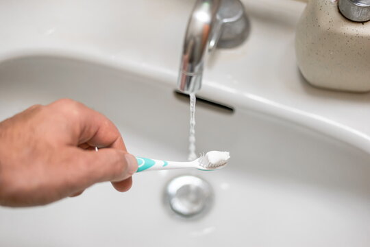 Male hand holding a toothbrush with white paste over bathroom sink and flowing tap water