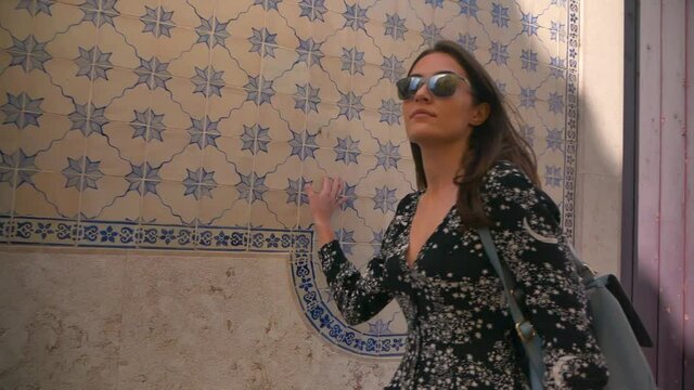 young happy caucasian tourist in glasses with backpack travels in portugal.girl walks down the street and touches traditional blue azulejo tiles on an old building in lisbon. woman enjoys architecture