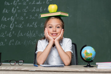 playful schoolgirl sitting at desk with book and apple on head