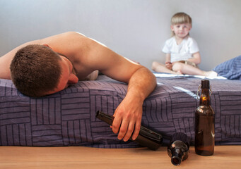 A drunken alcoholic man lies on the bed with a bottle of alcohol in his hand next to a small child....