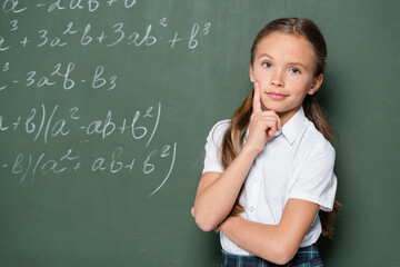 pensive schoolgirl with hand near face looking at camera near chalkboard with equations
