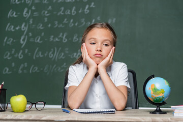 upset schoolkid sitting at desk near globe, notebook and apple