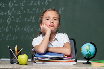 upset schoolgirl looking at camera near notebooks, globe and chalkboard on blurred background